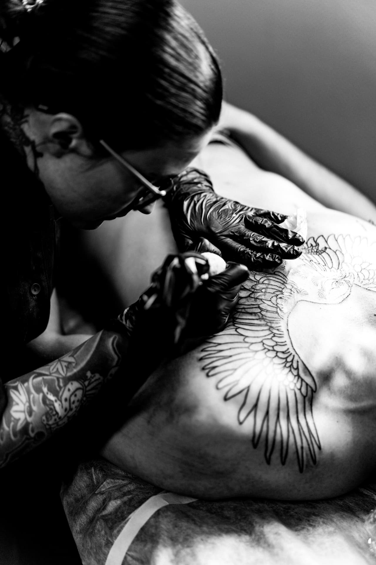 Tatoueuse penchée en séance, gants noirs et machine, traçant une aile sur le torse d'un client